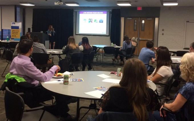Several people sit at tables in a room, watching a seminar.