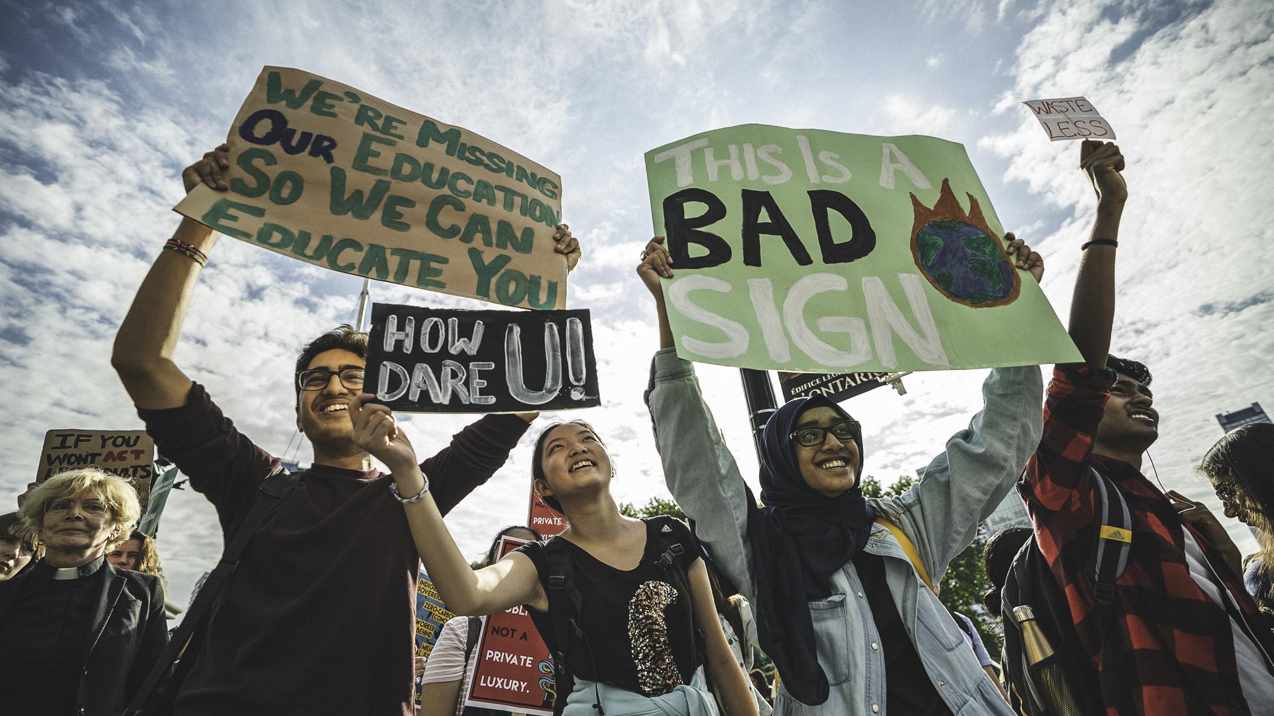 Three individuals smile holding up their signs, reading: "We're Missing Our Education So We Can Educate You," "How Dare U!" and "This Is A Bad Sign."