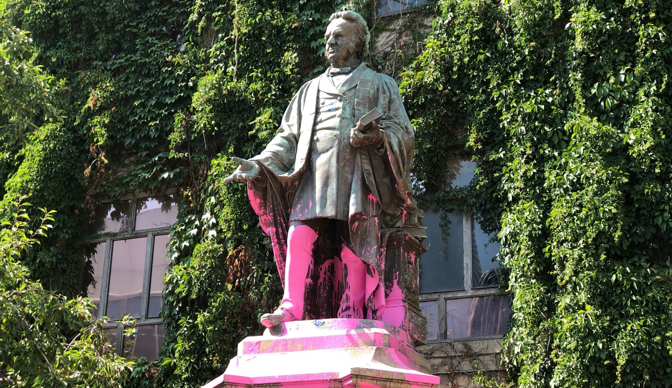 A tall statue of Egerton Ryerson stands against a green, mossy wall. The bottom of the statue has been splashed with pink paint.
