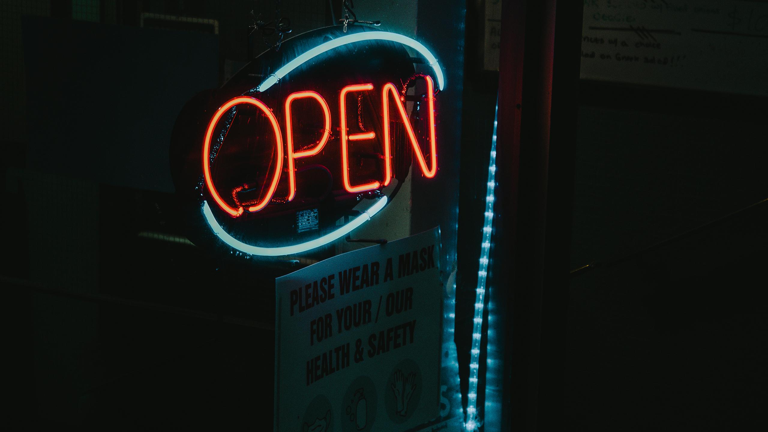 A picture of a neon open sign. Underneath, a smaller white cardboard sign says "please wear a mask for your/our health & safety."