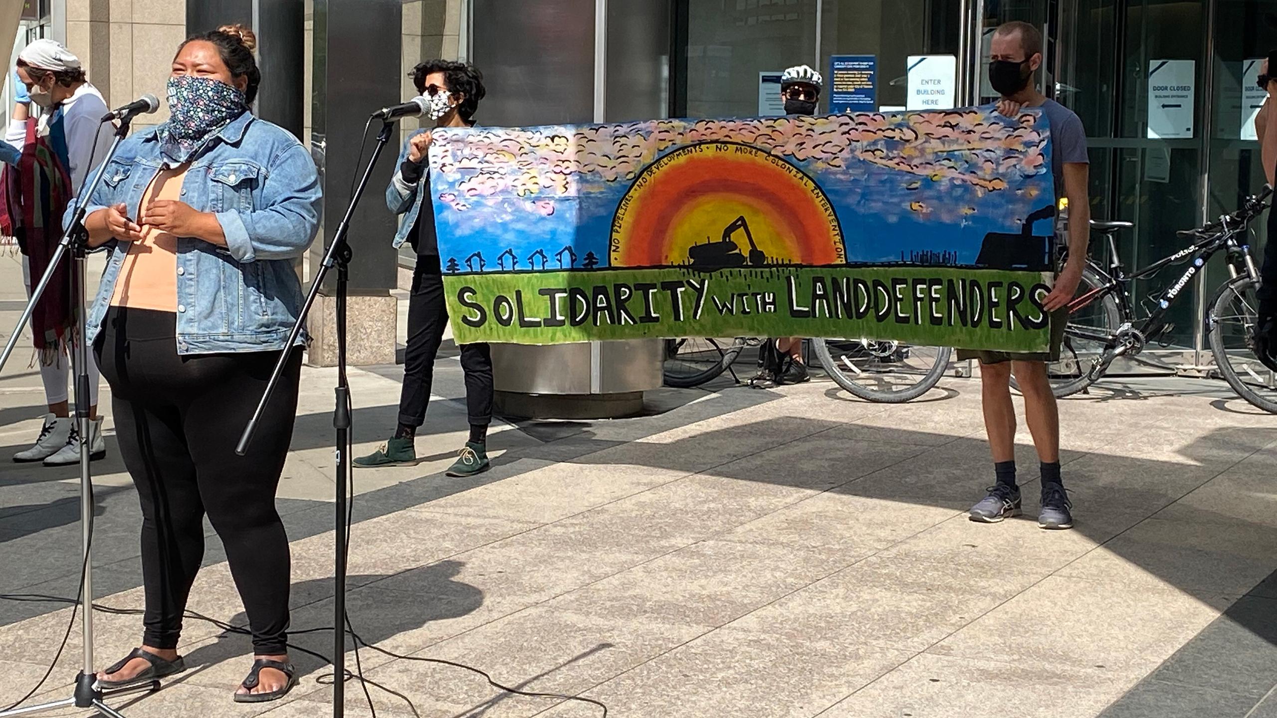 Photo of a person wearing a bandana over their face speaking into a microphone outside. Behind them, there are two people holding a banner with a painting of a sunset and text that says "solidarity with land defenders."