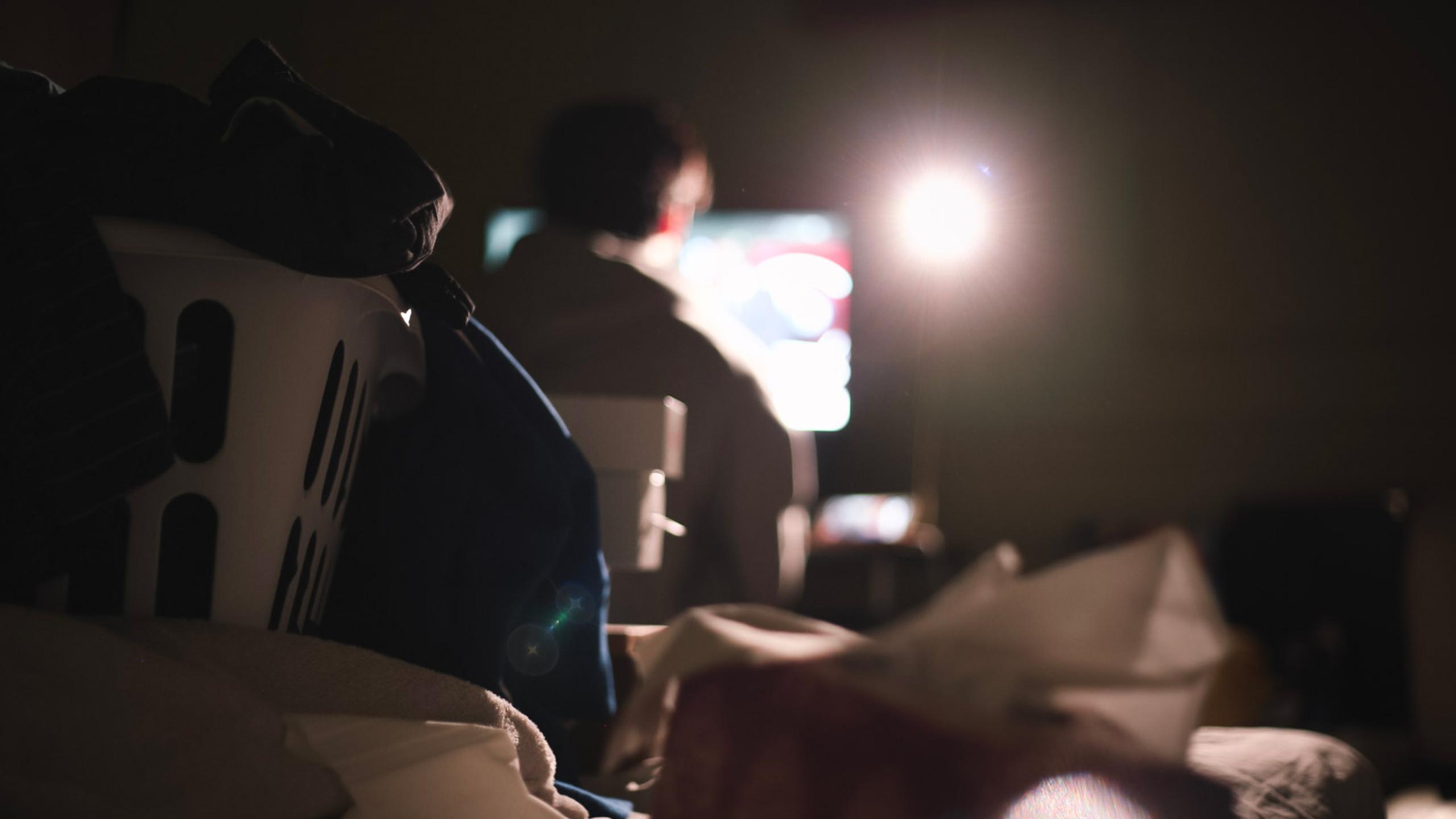 Photo of a student sitting at his desk looking at a computer. His room is messy, with laundry and tissues everywhere.