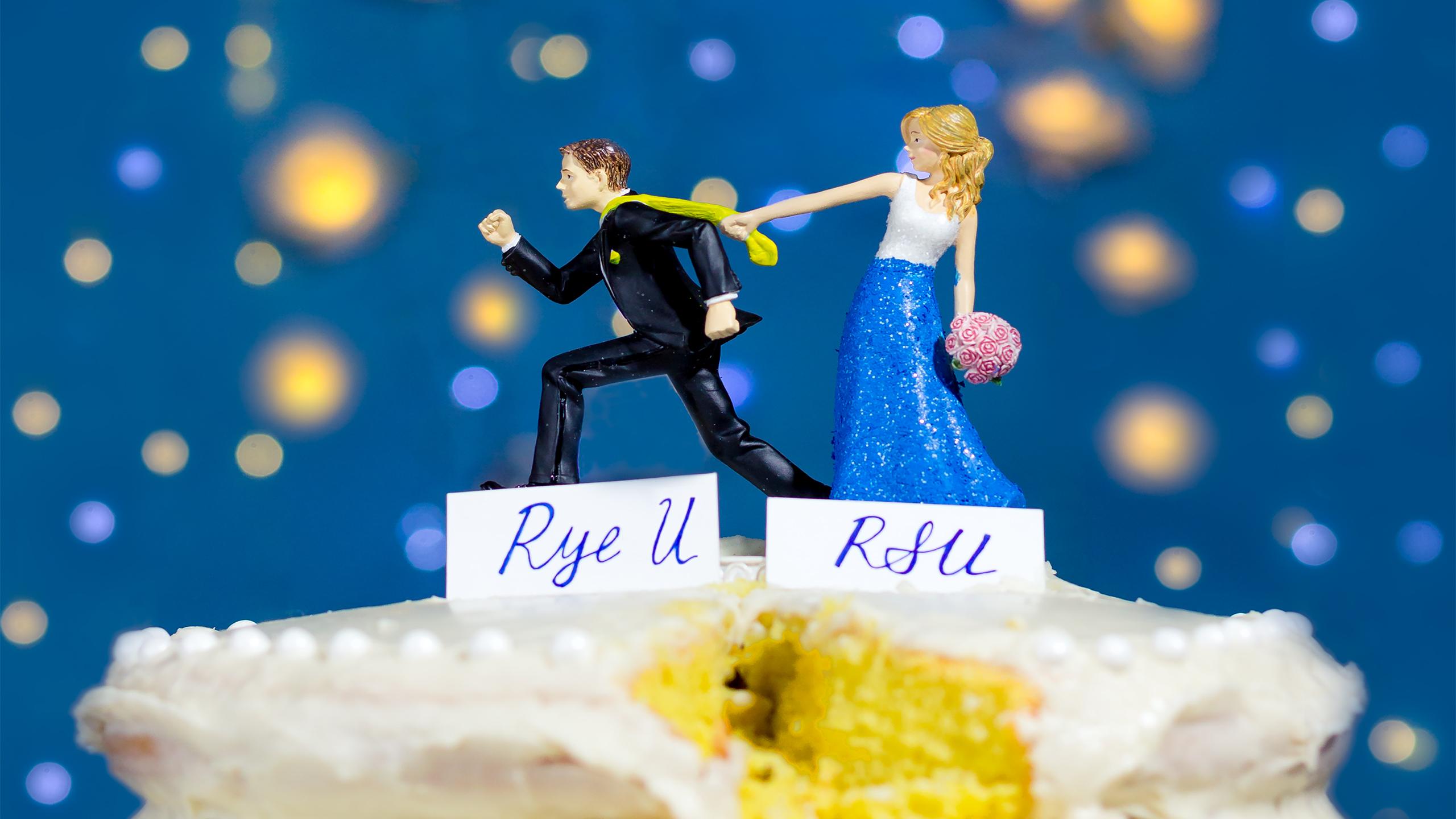Wedding cake toppers showing a bride holding onto her groom's tie as he tries to run away. A nameplate under the bride says RSU and under the groom says Rye U.
