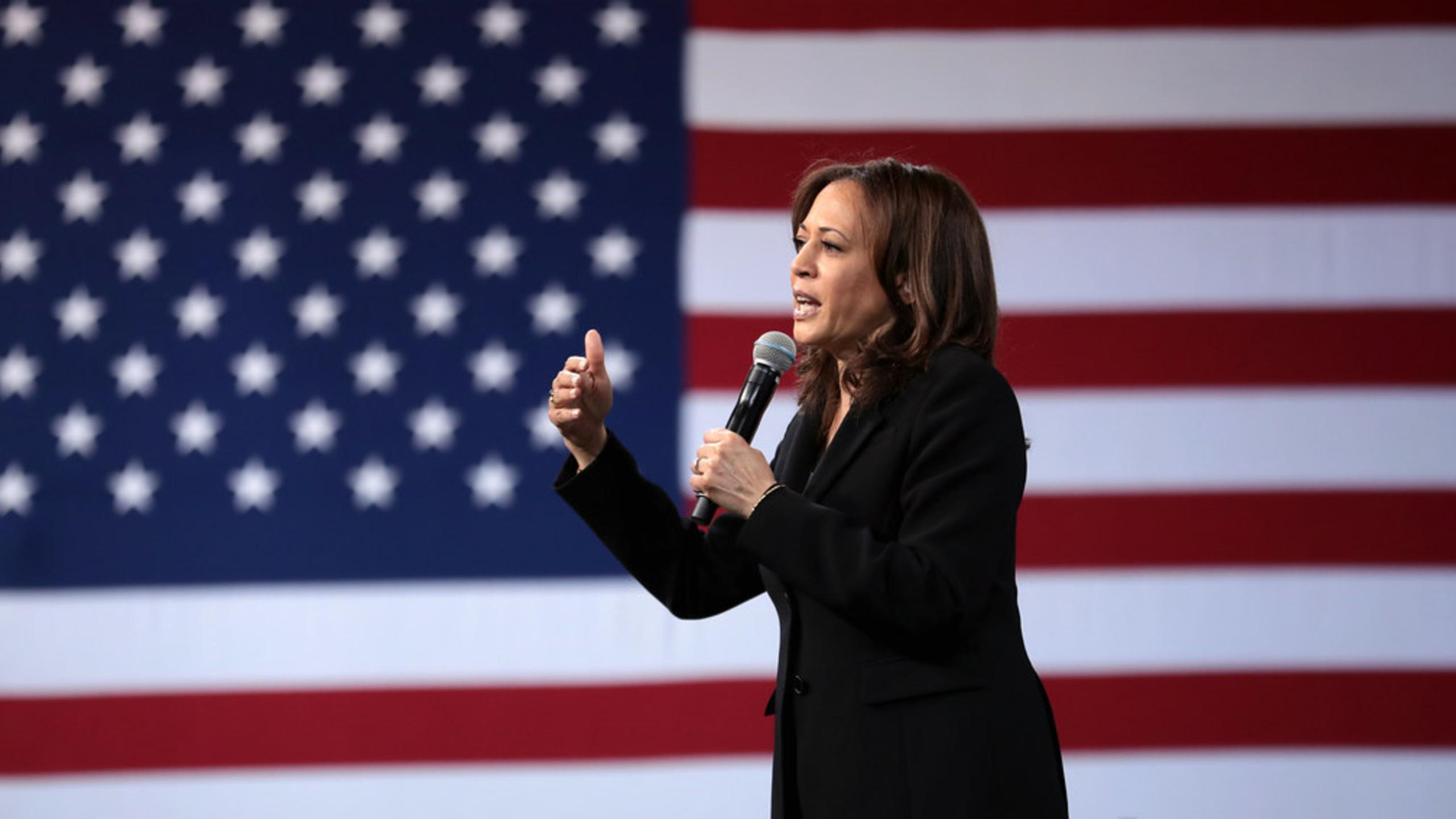 Kamala Harris wearing a black suit holding a microphone in front of an American flag