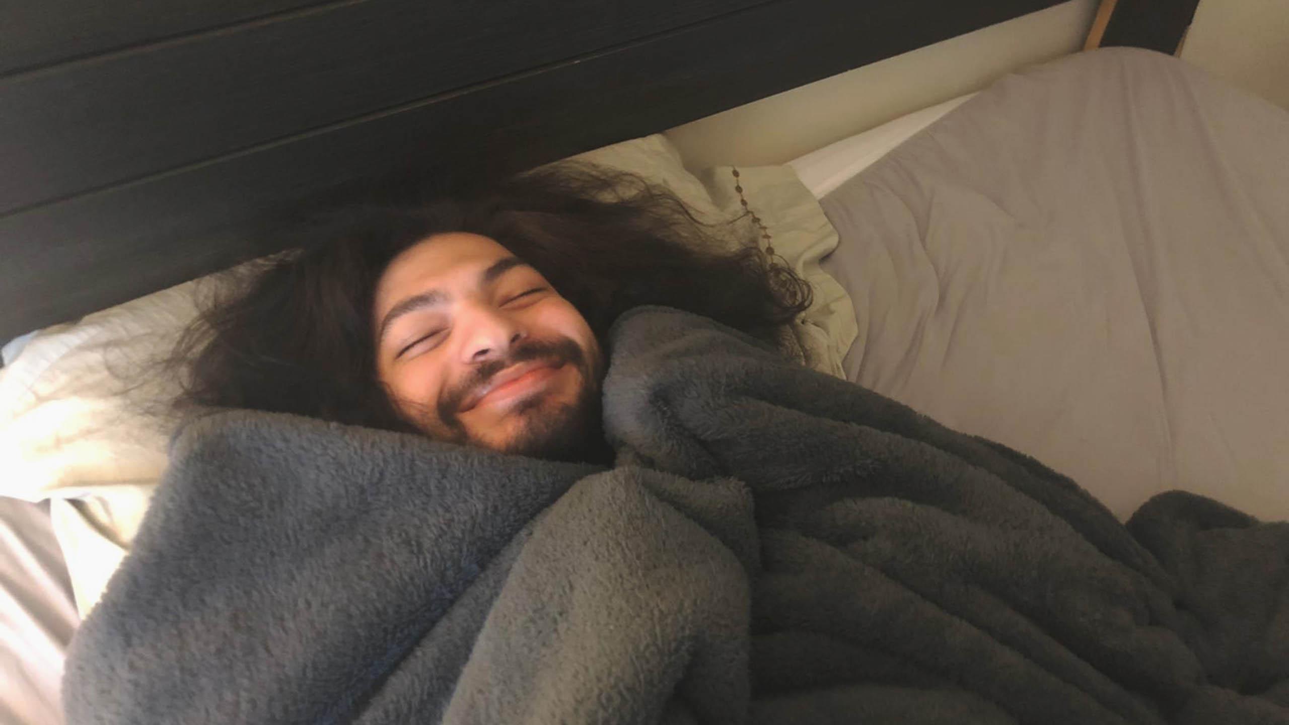 Man laying on bed, covered with blanket, smiling happily.
