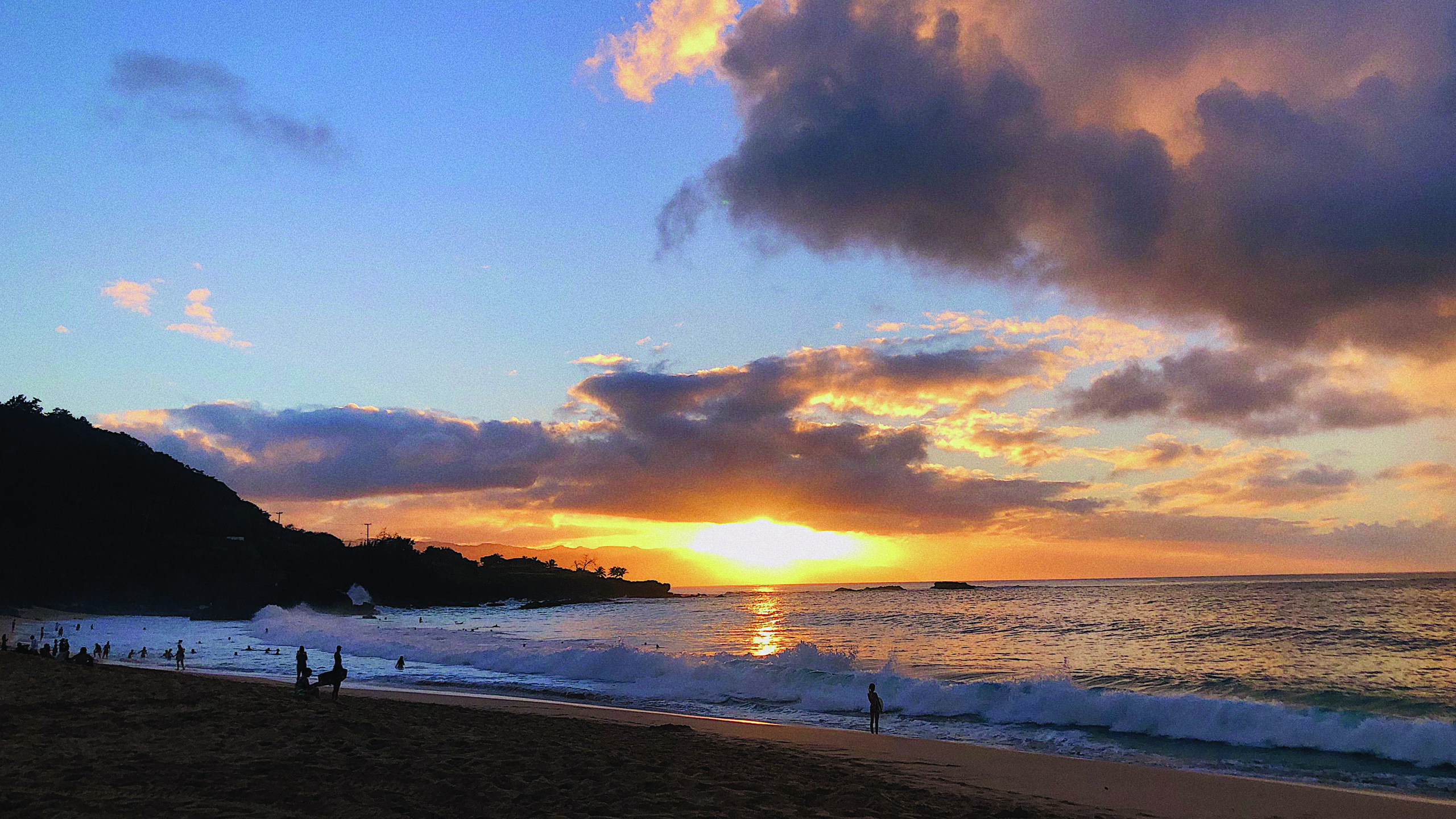 a landscape photo of a vibrant sunset over a beach, with a blue sky, few clouds and the sun touching the horizon over the water. The bright yellow sun reflects on the water.