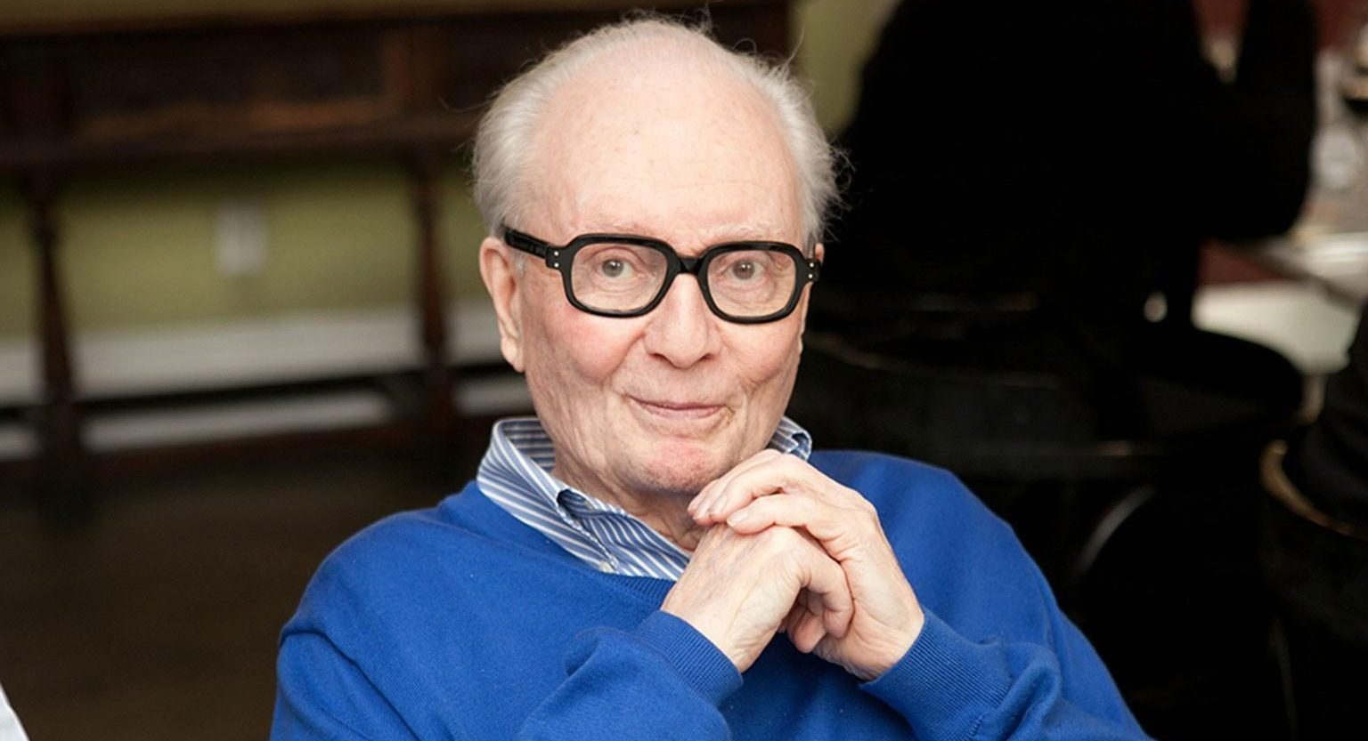 An older man with black rimmed glasses and a blue sweater sits and smiles
