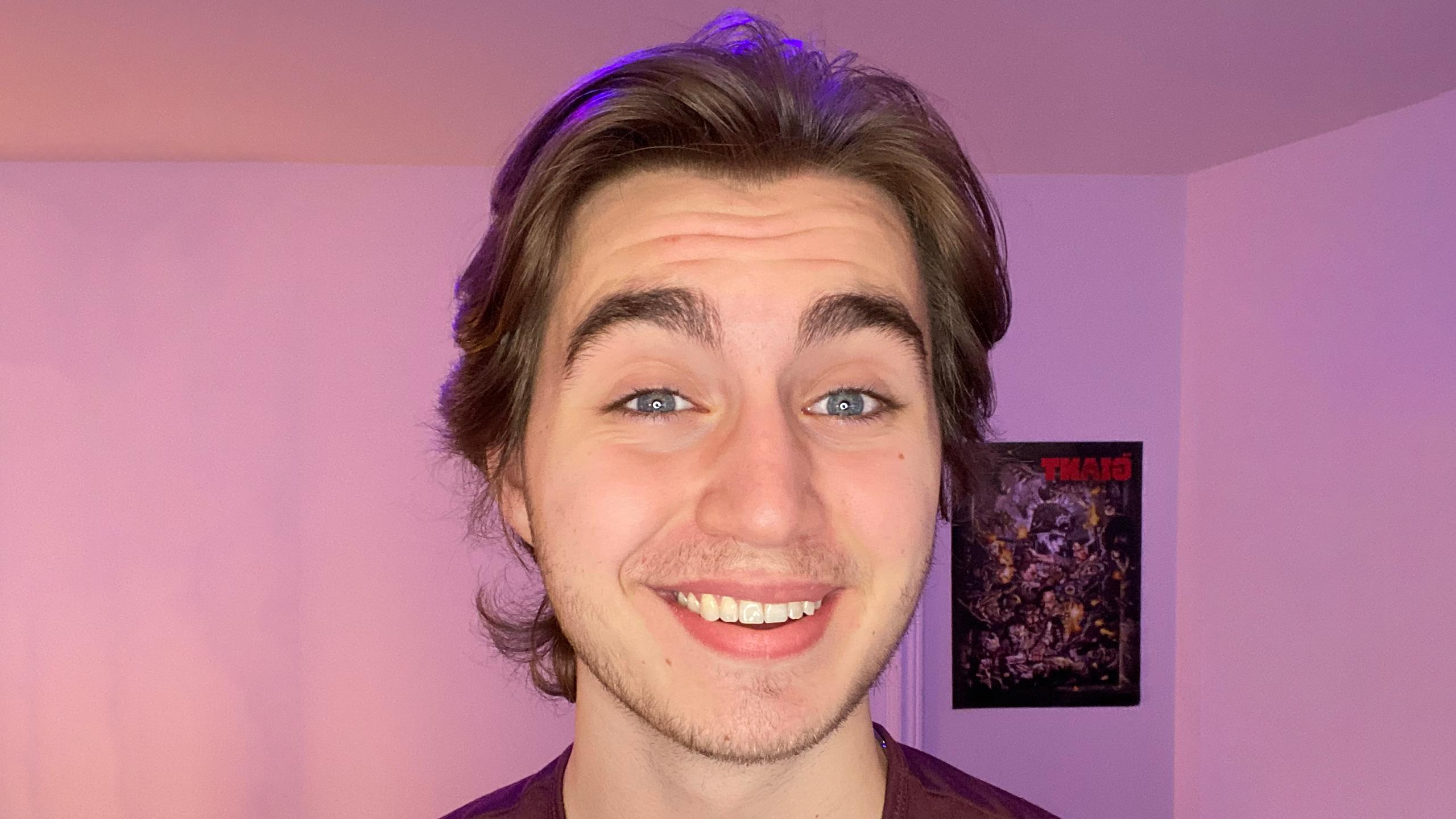 A picture of Michael DiMuccio, a young white man with medium-length light brown hair. He's raising his eyebrows and smiling. The background behind him is an empty wall other than a poster and pink lighting.