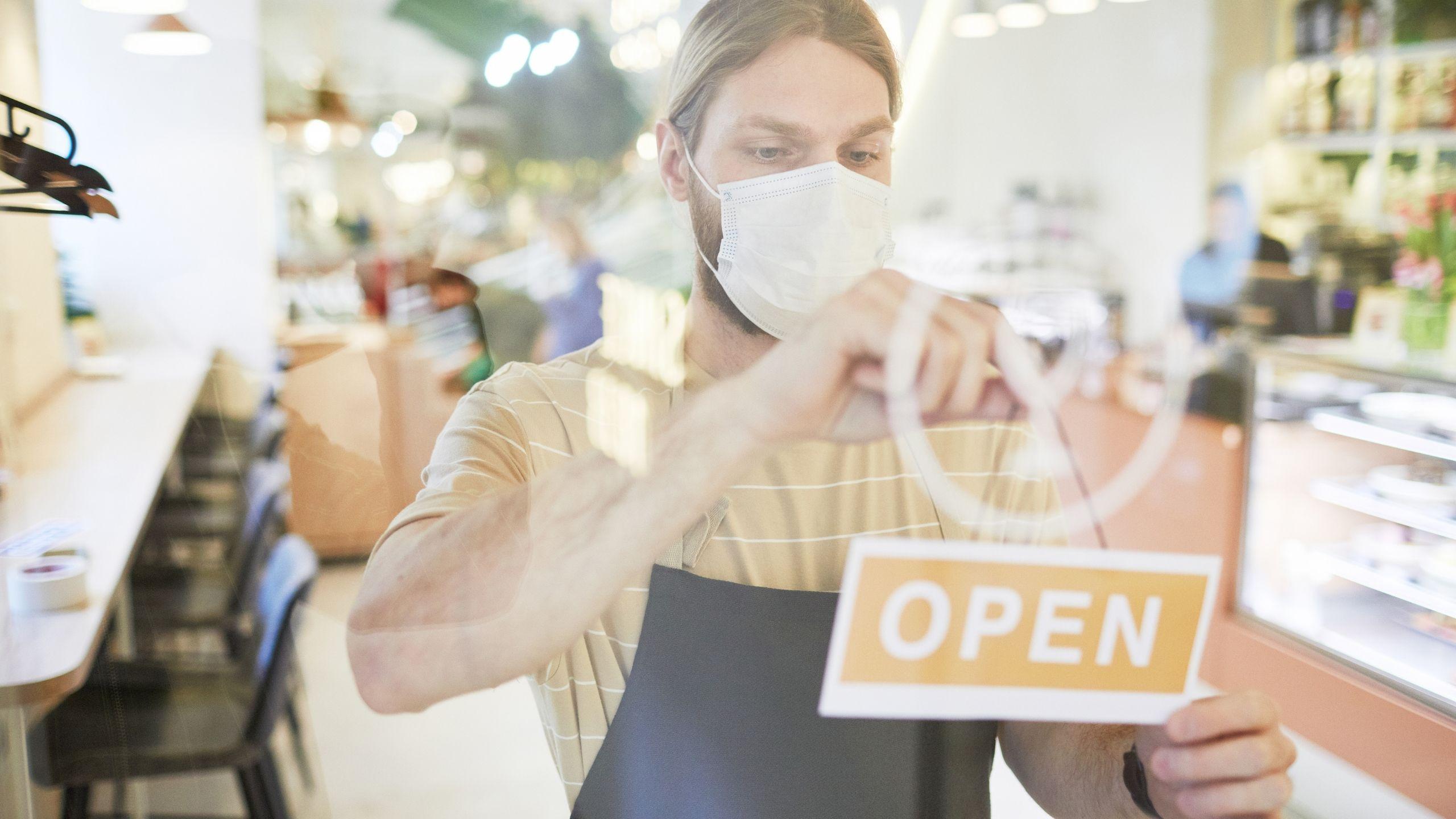 Person wearing a mask while attaching an open sign on the storefront made of glass.