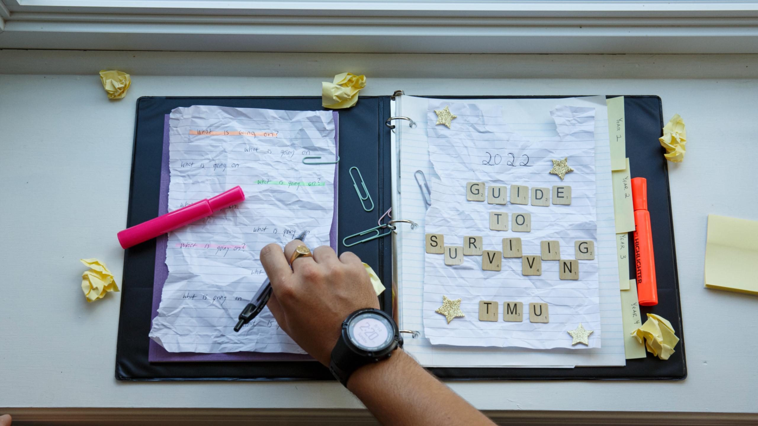 Open binder with two crumpled pages, page on the left has the words "2022 guide to surviving tmu". Page on the left have a pink highlighter across and the words "what is going on". Arm inserted from the bottom and is writing on the left page. Orange highlighter on right side of the binder. Crumpled up yellow paper around the binder, paper clips littered in centre of binder.