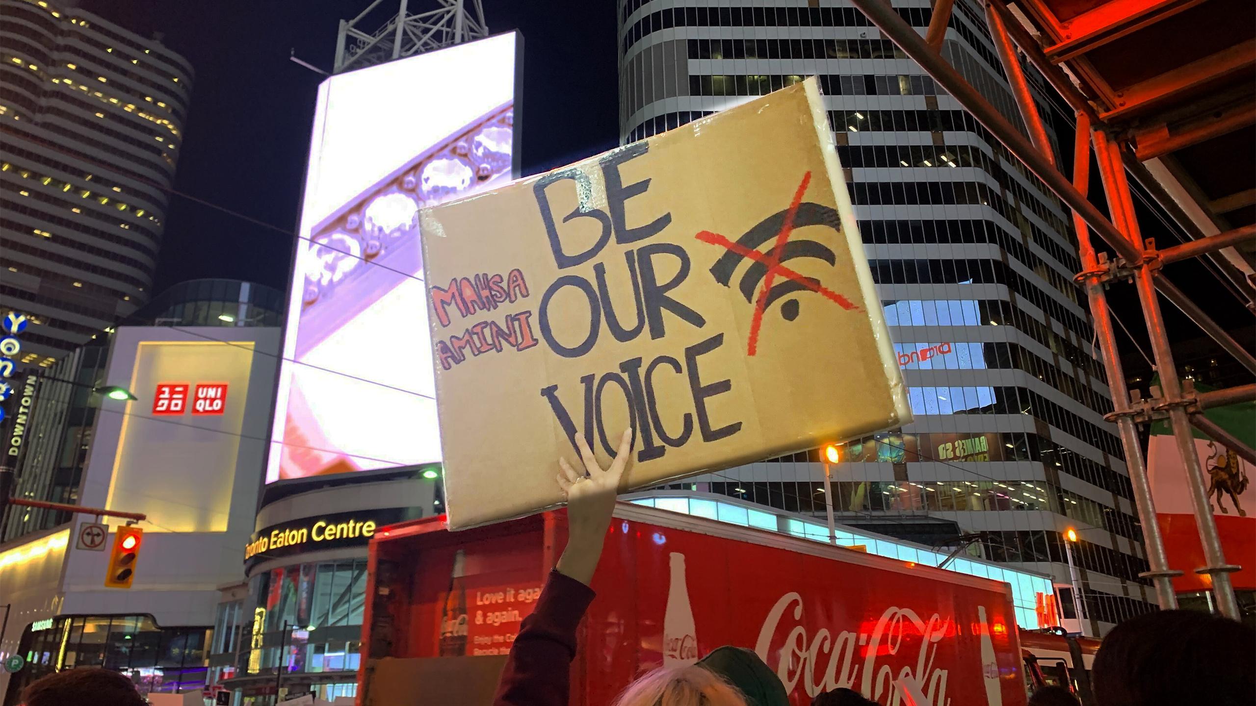 A protestor holding a sign at Yonge-Dundas Square that reads "Mahsa Amini, Be Our Voice"