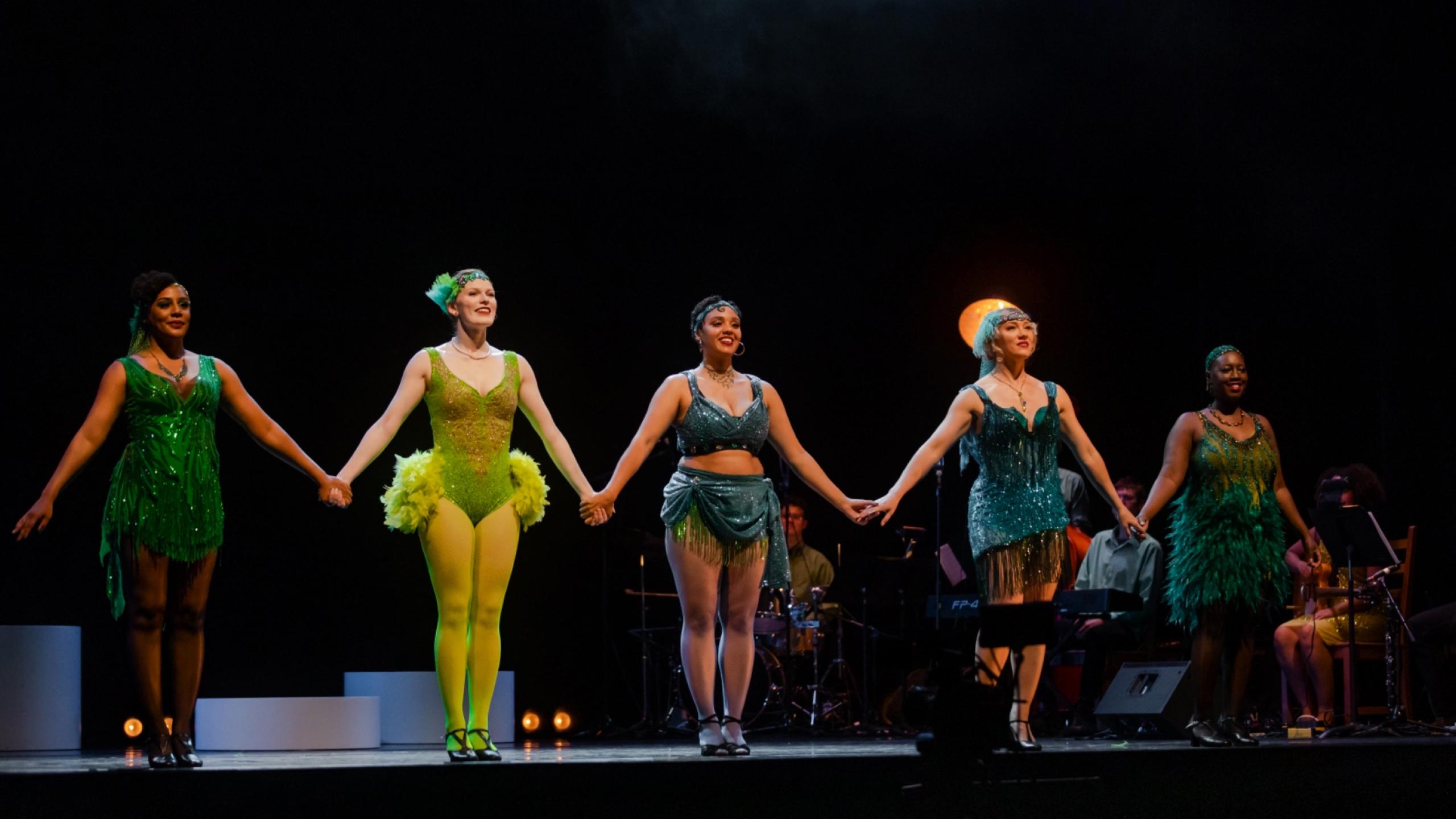 A photo of five women on stage holding hands and speaking to the crowd in 1920s attire