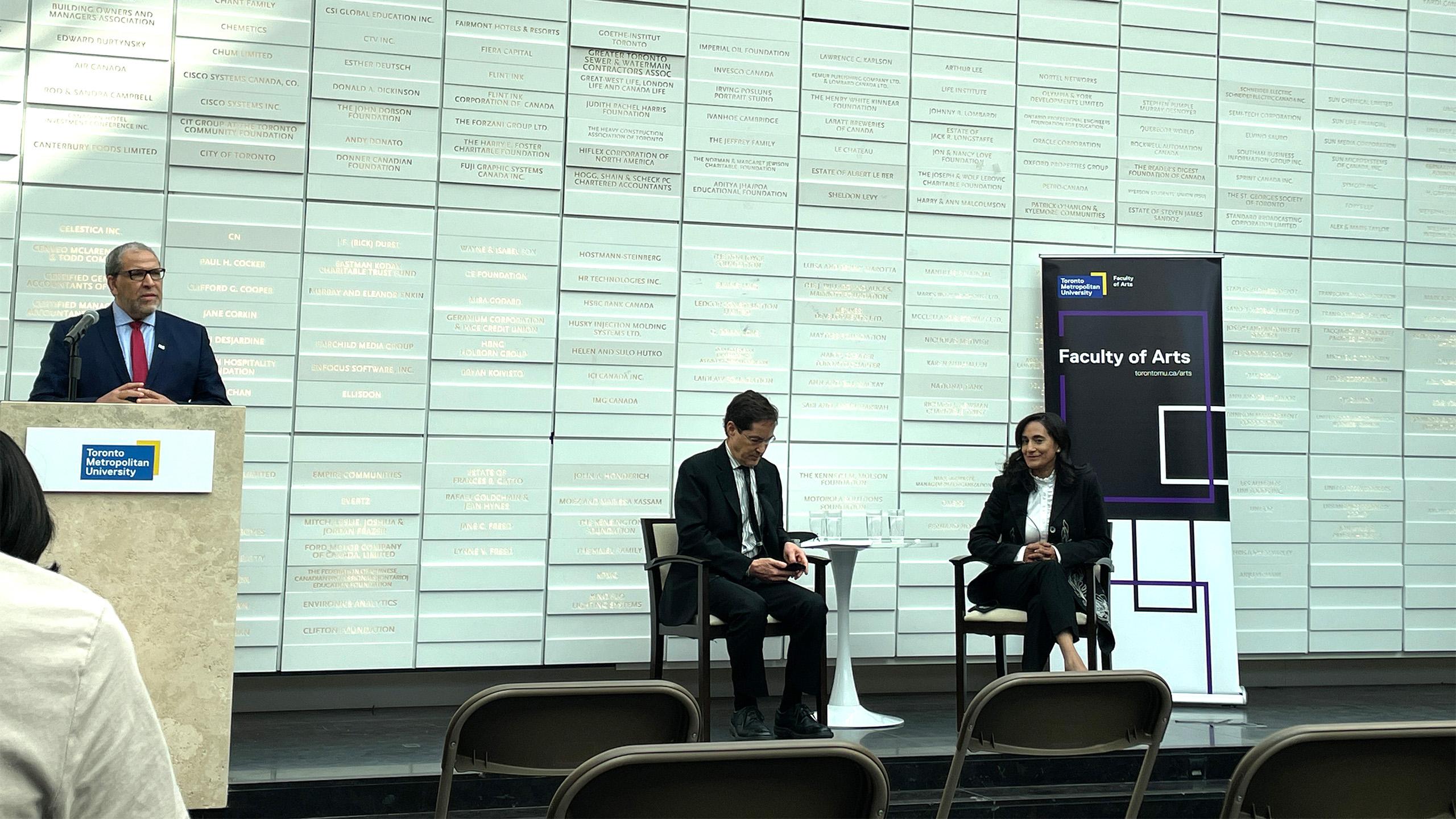 to the left two people are sitting on a stage looking at the speaker to the right at a podium