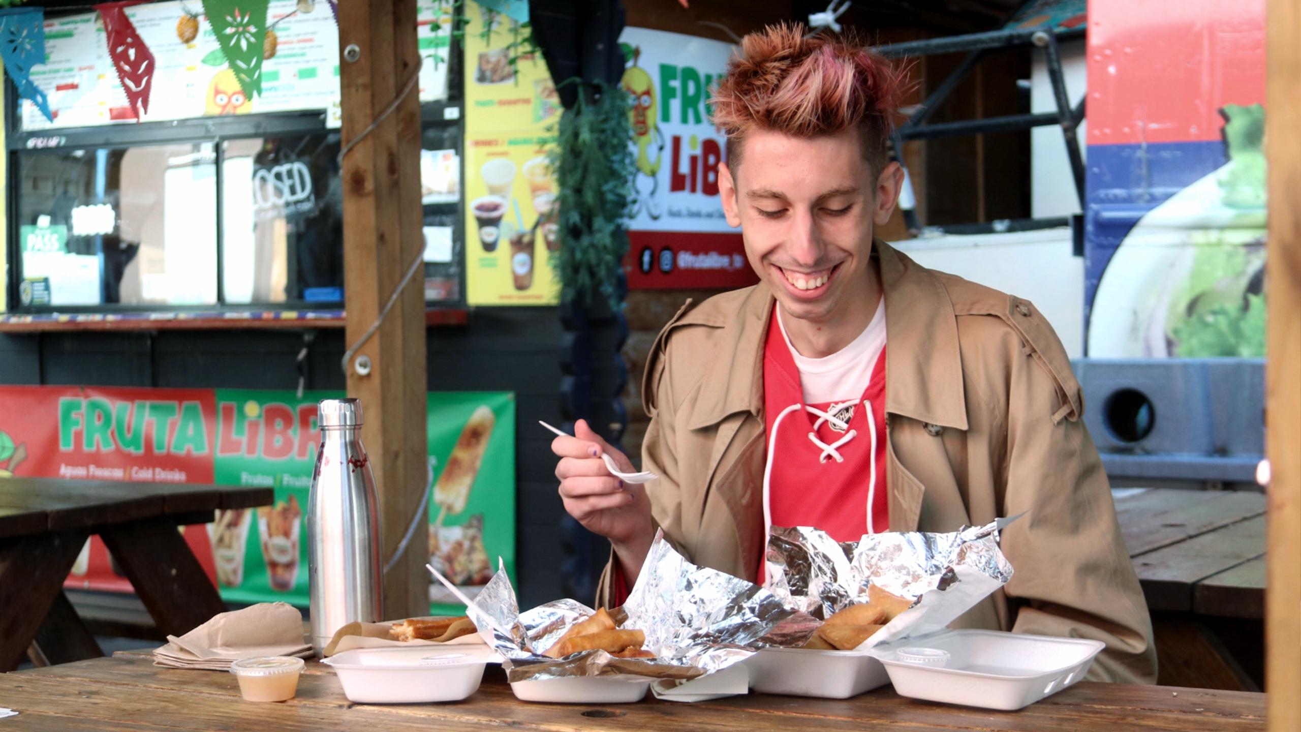 A person at a picnic table in the food market across from the SLC smiling at the plates of food in front of them