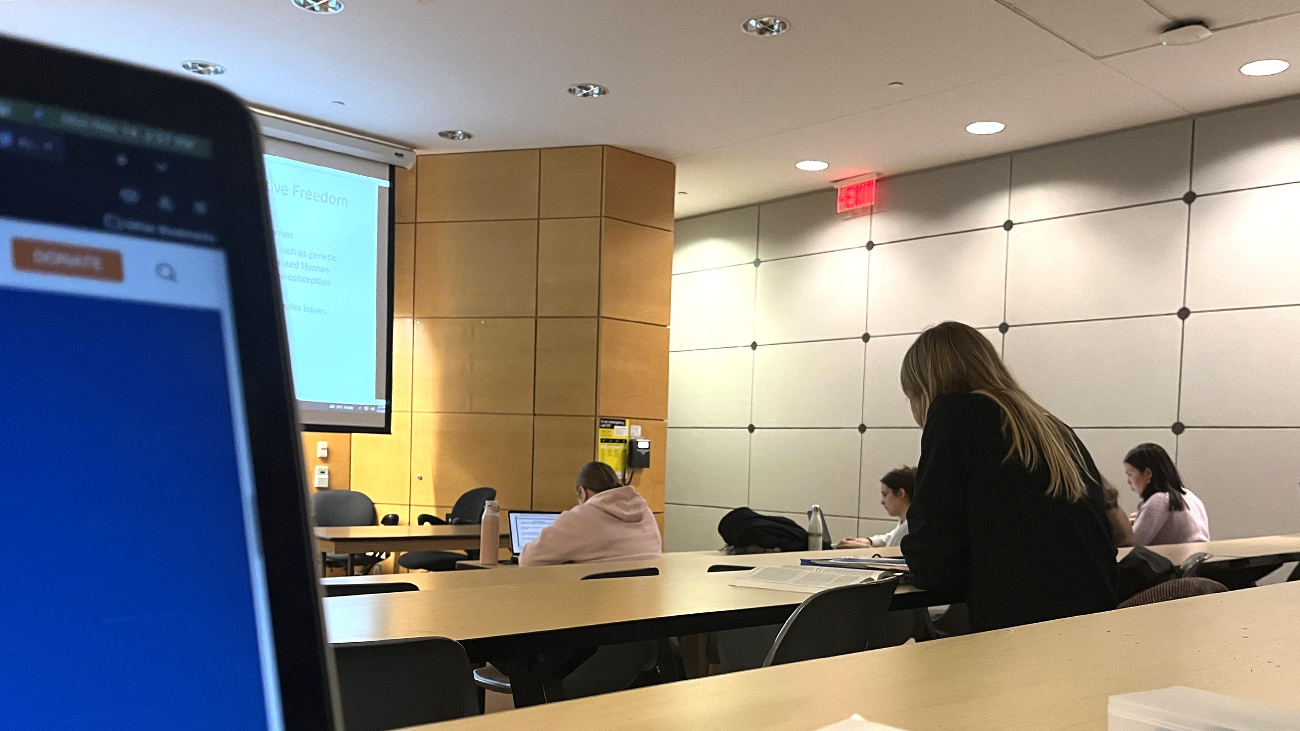 In the left side, a laptop screen is visible. The rest of the image shows a lecture hall with students seated around the desks taking notes.