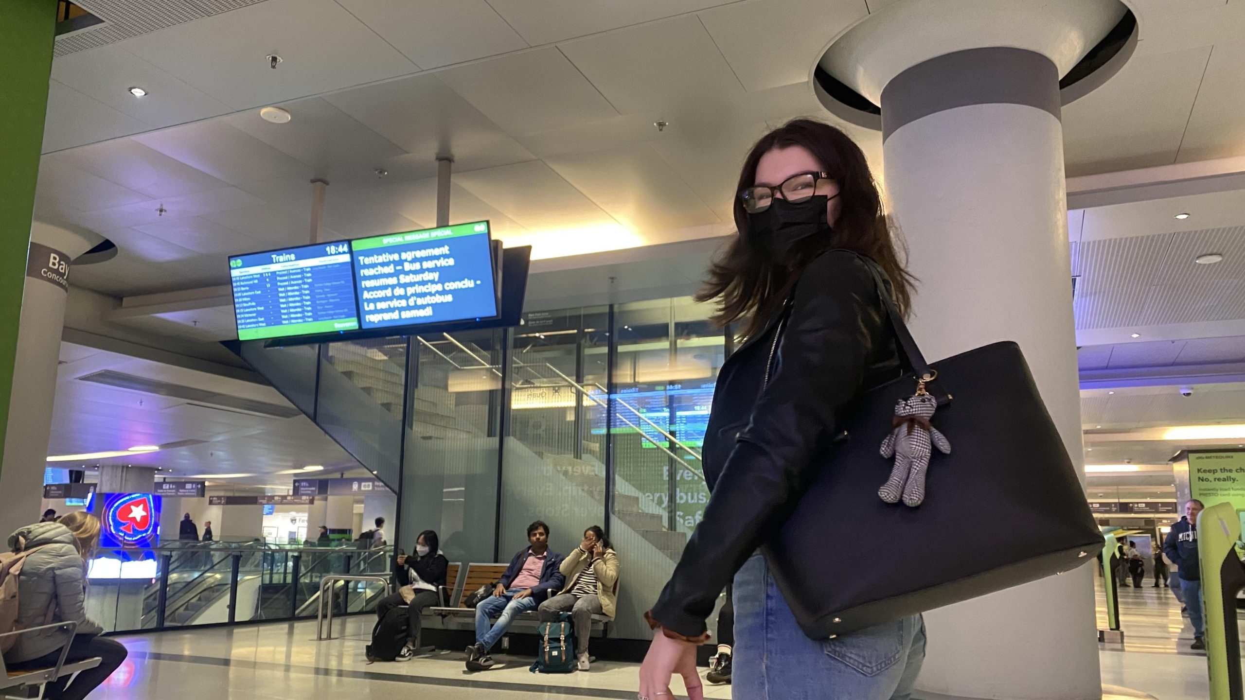 A student with dark hair and a black jacket waits un Union Station to take GO Transit. Behind her is a TV screen displaying the departure times of trains.
