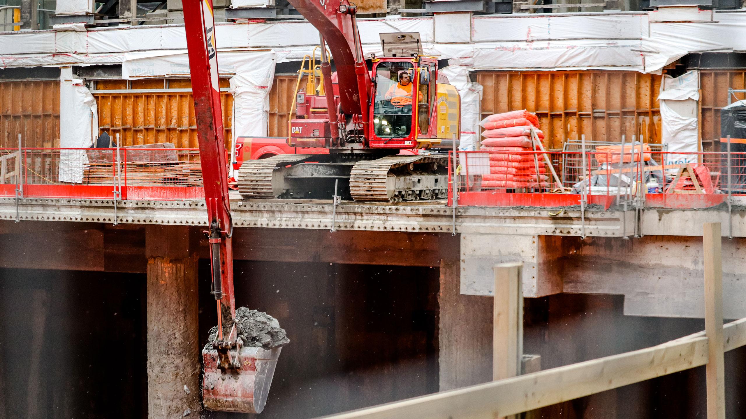 A man wearing an orange vest maneuvers a red excavator at a fenced construction site
