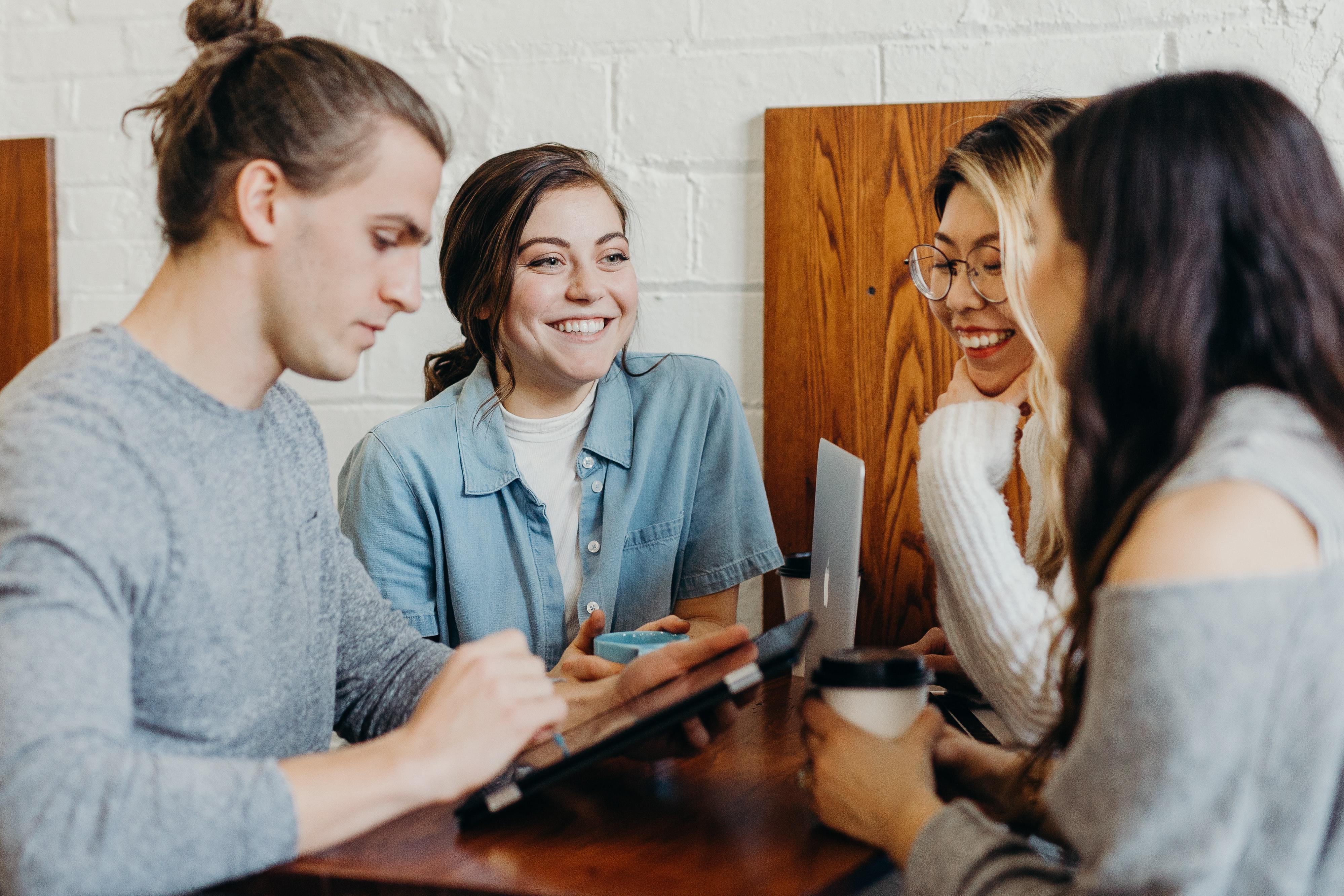 Four people sit at a wooden table using various electronic devices, drinking coffee and smiling