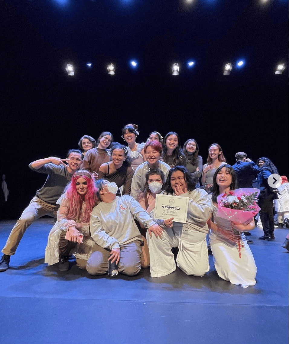 A group of a cappella performers kneel on stage. One member holds a certificate while the other holds a flower bouquet.