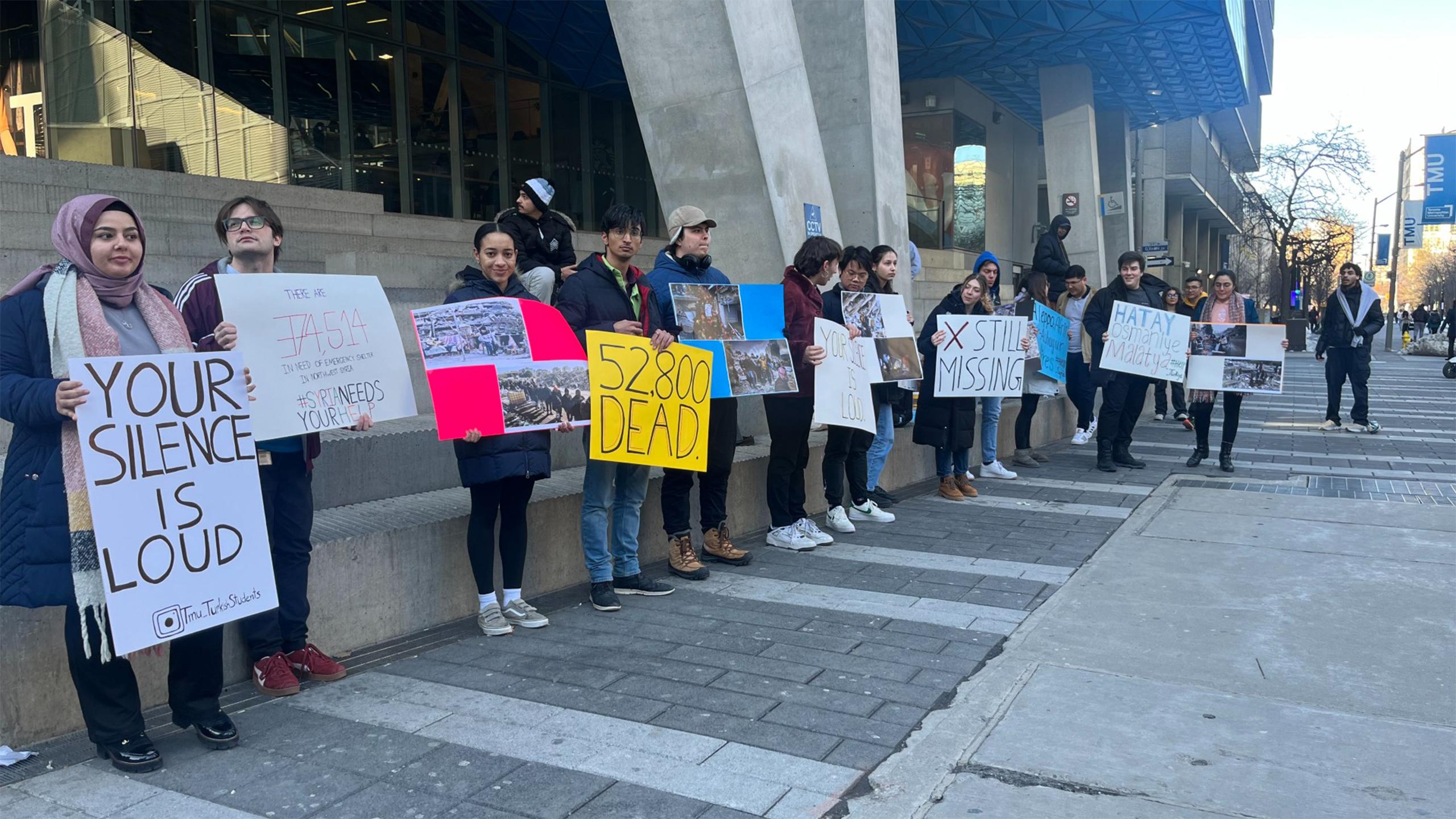 A line of dozens of students in front of the SLC holding signs reading "your silence is loud" and "52,800 dead"