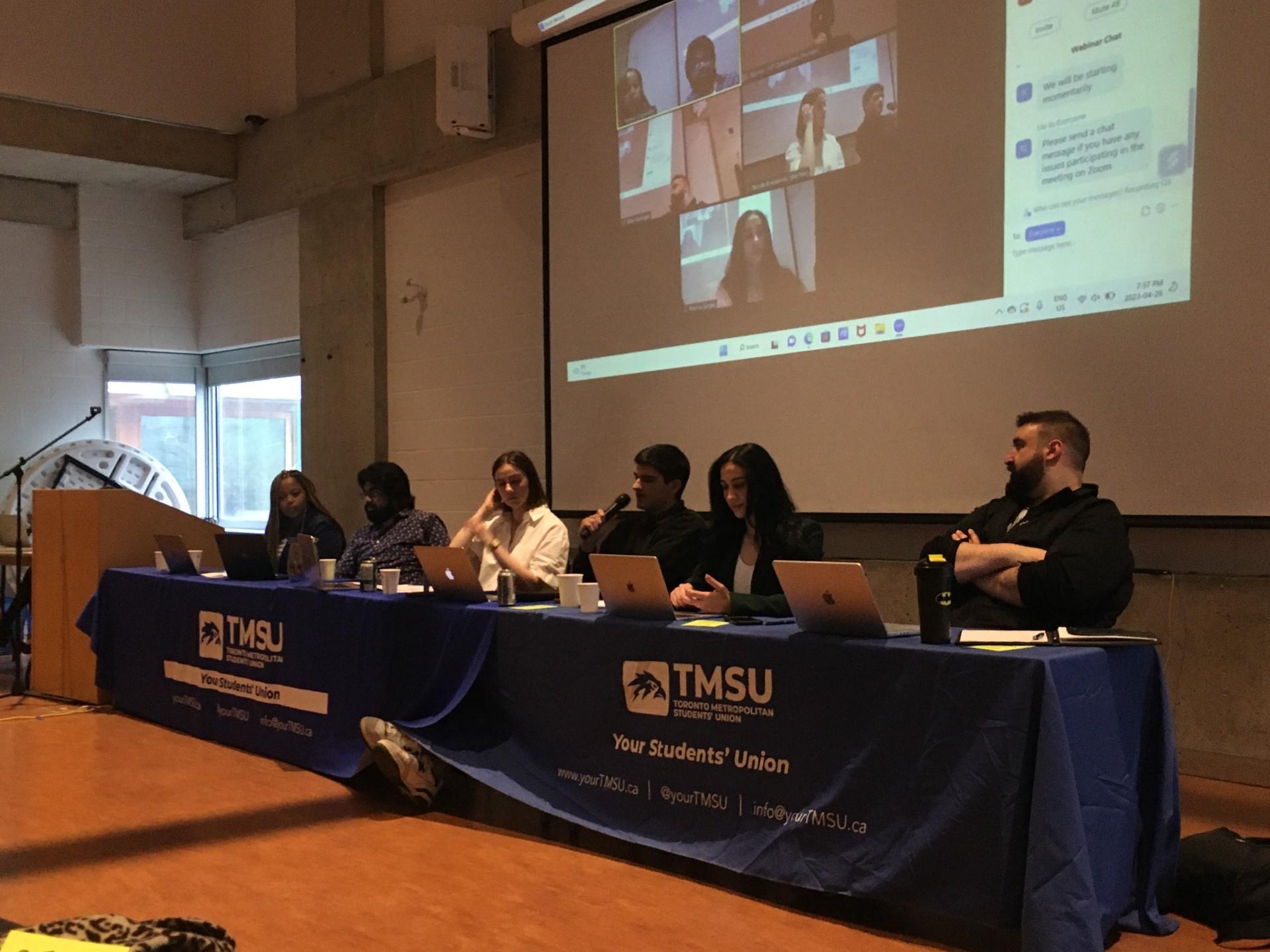 Six people sit at table with TMSU tablecloth. Zoom screen behind them.