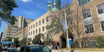 A sunny summer day on Gould Street outside of Kerr Hall. A group of orientation attendees stand outside of a gate while a row of cars appears in the foreground.
