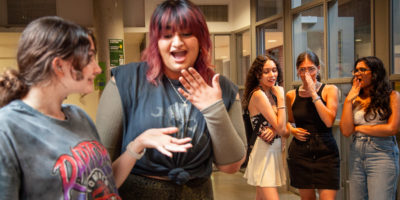 Two students laugh together while three more students behind them whisper to one another in the hallway