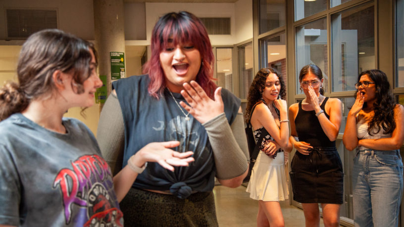 Two students laugh together while three more students behind them whisper to one another in the hallway