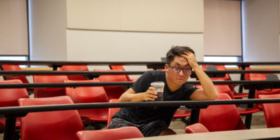tired-looking student sits alone in a classroom at a desk holding a coffee cup in one hand and holding his forehead in the other hand propped up on the desk