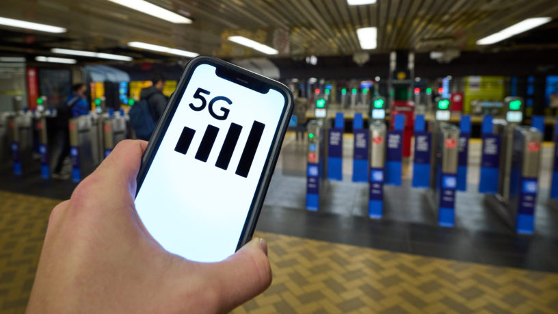 A person holds a phone with a 5G symbol on it's screen while in front of ticket gates in a TTC subway station