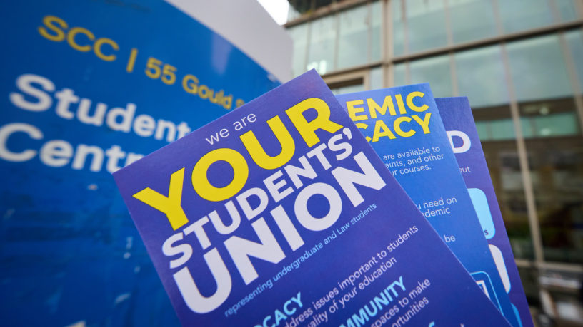 Flyers in blue, yellow and white being held up in front of SCC building sign
