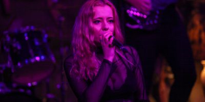 Amy Kisser sings into a microphone, while standing in front of a drummer and bass guitarist. The entire scene is illuminated with pink lighting.