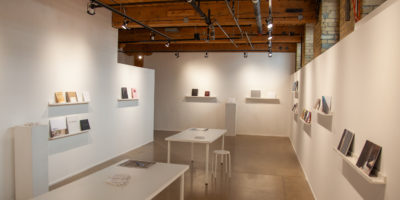 A white room with wood-paneled ceilings has walls scattered with shelves that hold various photobooks. There are two white tables in the middle of the room with stools and one book each.