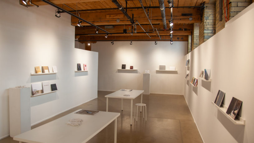A white room with wood-paneled ceilings has walls scattered with shelves that hold various photobooks. There are two white tables in the middle of the room with stools and one book each.