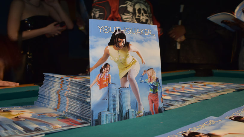 A stack of Youthquaker magazines lays atop a billiards table with one of the magazines lays propped against the stack, showcasing its cover of a woman towering over skyscrapers.