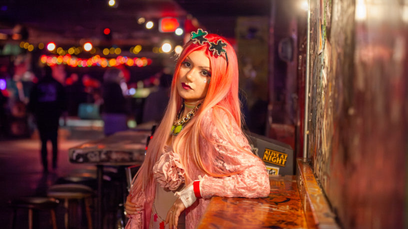 Carina leans against a counter while looking into the camera. The Sneaky Dee's stage can be seen illuminated in the background.