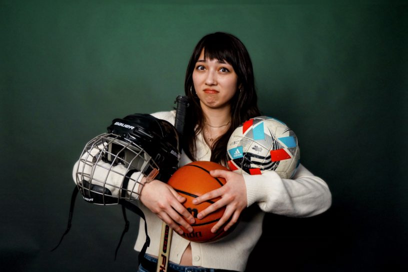 A confused girl holding up a helmet, basketball, hockey stick and soccer ball.