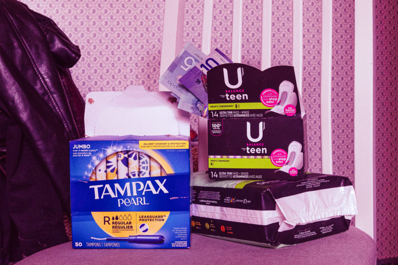 A stack of pads and tampons sit on a table with cash peeking out of one of the pad boxes.