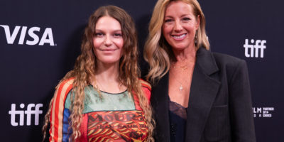 Two, well dressed women with long hair smiling at the camera in front of a black backdrop with the words "TIFF" and "VISA" on it.