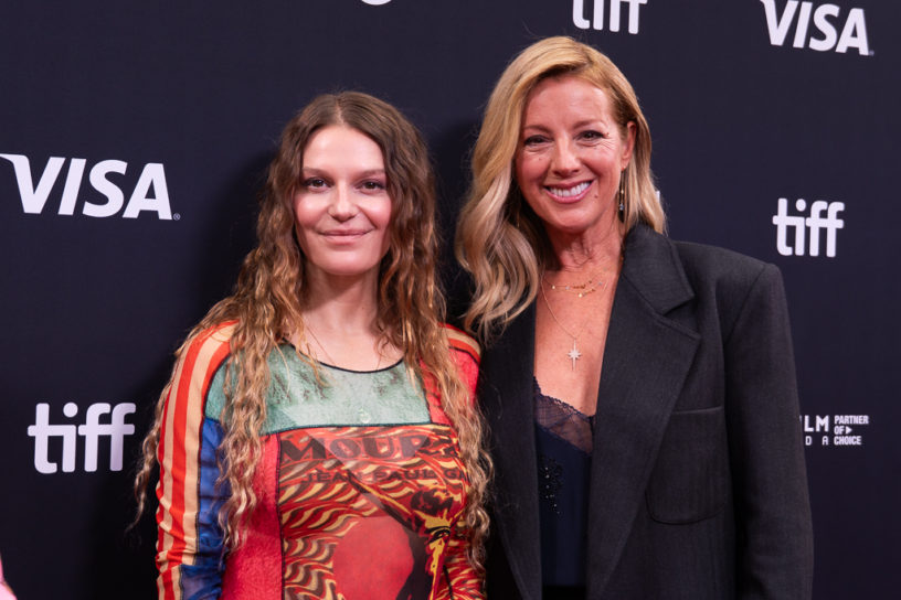 Two, well dressed women with long hair smiling at the camera in front of a black backdrop with the words "TIFF" and "VISA" on it.