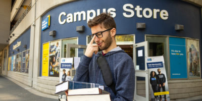 Student holding textbooks and pushing up their glasses in front of the Campus Store