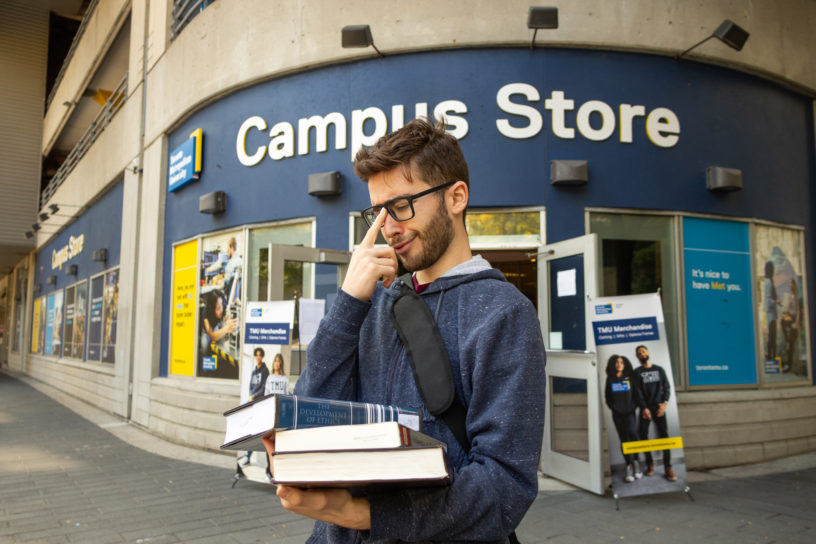 Student holding textbooks and pushing up their glasses in front of the Campus Store