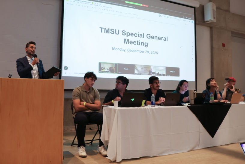 Photo of six people sitting at a white table in front of a powerpoint on a large screen that says "TMSU Special General Meeting."