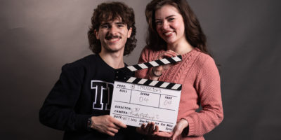 Two people look into the camera smiling while they hold a clapperboard.