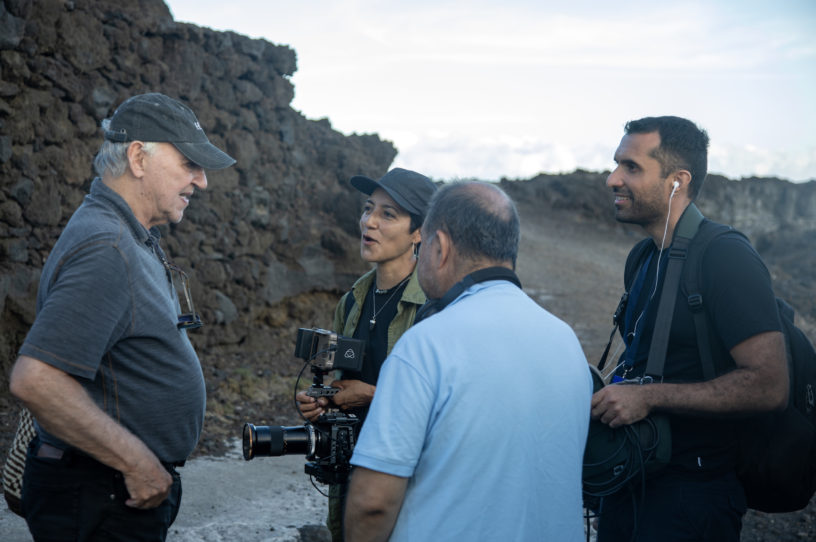 Four people standing in a circle on a rocky hill with camera equipment, smiling and talking to each other.