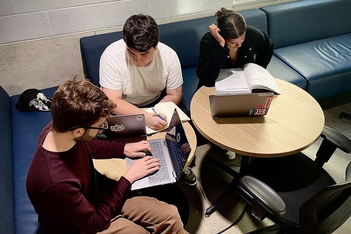 Three students sitting at a table studying with laptops and books open.
