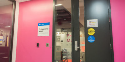 Image of the outside of a room with a pink wall with a sign titled "professional music student lounge"