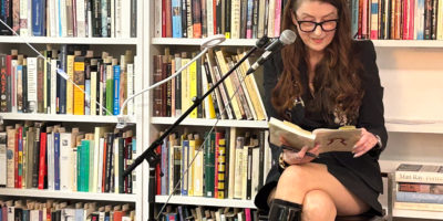 Image of a person looking down at a book reading into a microphone, sitting in front of a bookshelf.