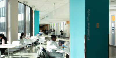 Image of people studying at tables in a building with text on a blue pillar that reads "TRSMRetail #PopUp."