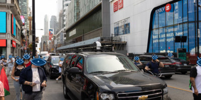 Photo of a busy street with a facial recognition camera on top of a black SUV, there are illustrations of blue eyes over every persons face.